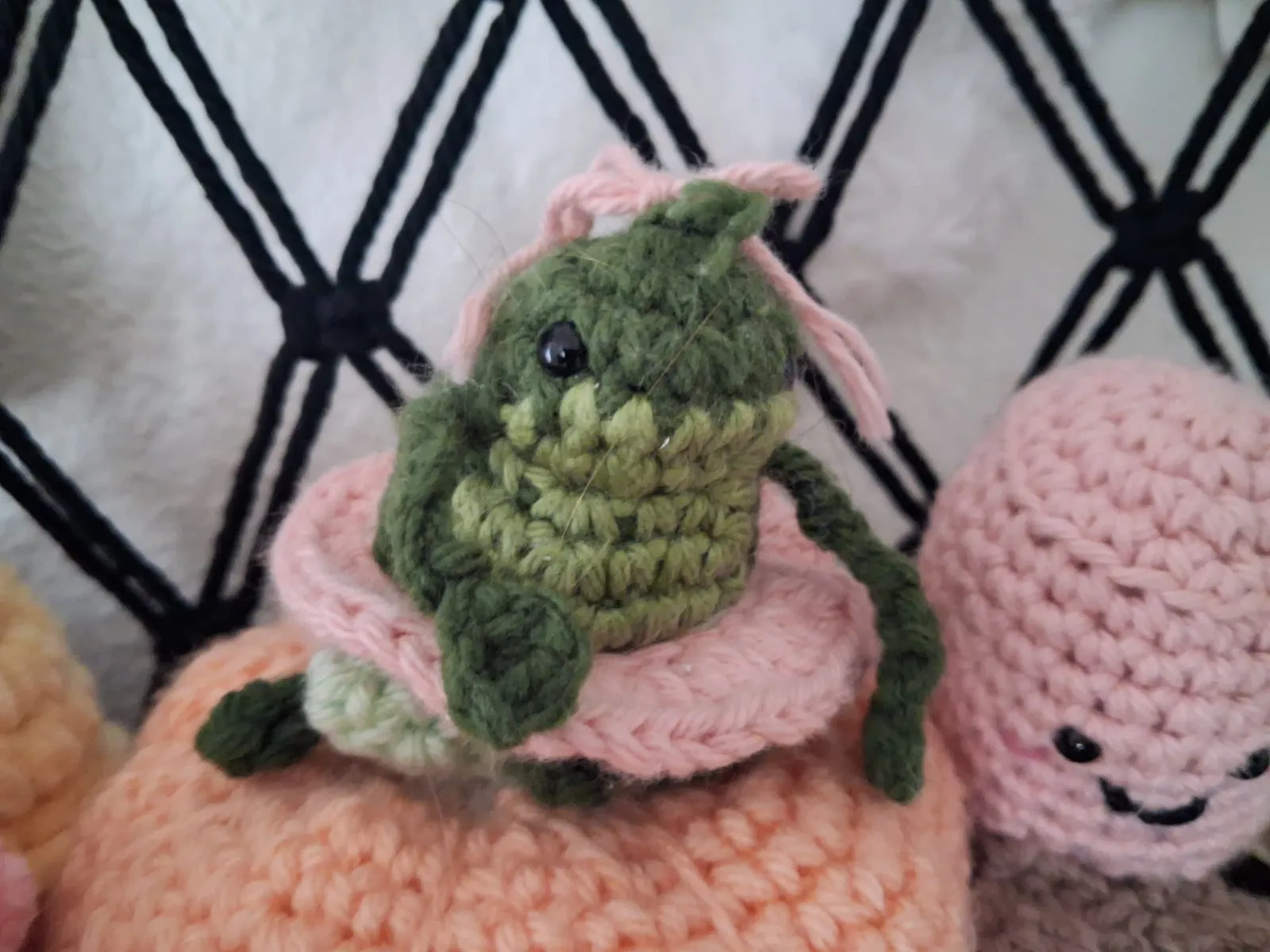 A small green crochet frog wearing a pink lily-pad hat with a pink bow